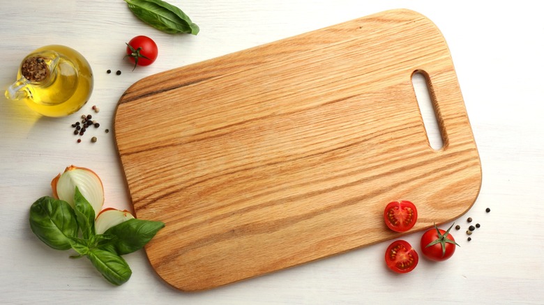 Wooden cutting board on white surface next to tomatoes, basil, and bottle of oil