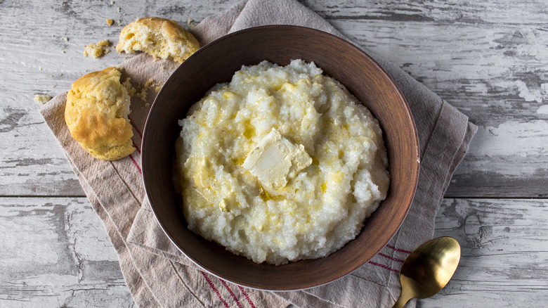 a bowl of buttered grits set on a napkin with a spoon and biscuit beside it