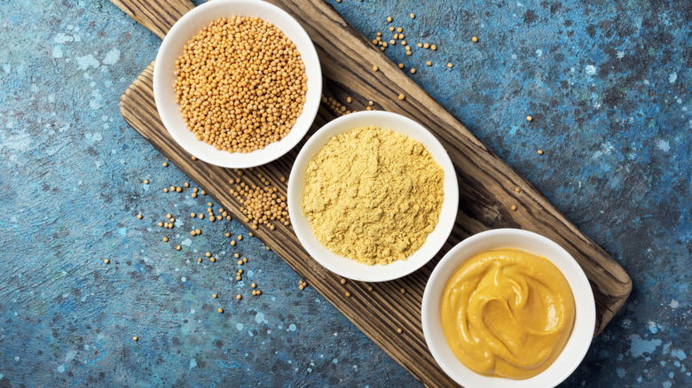 Mustard seeds, mustard powder, and prepared mustard in white bowls on cutting board
