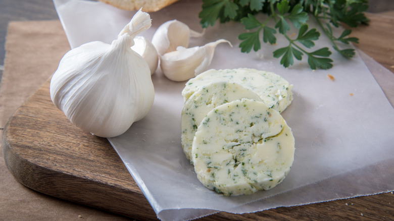 Rounds of sliced garlic butter on parchment, beside whole cloves of garlic and parsley