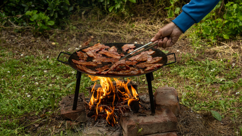 a person tends meat cooking over an open wood fire outdoors