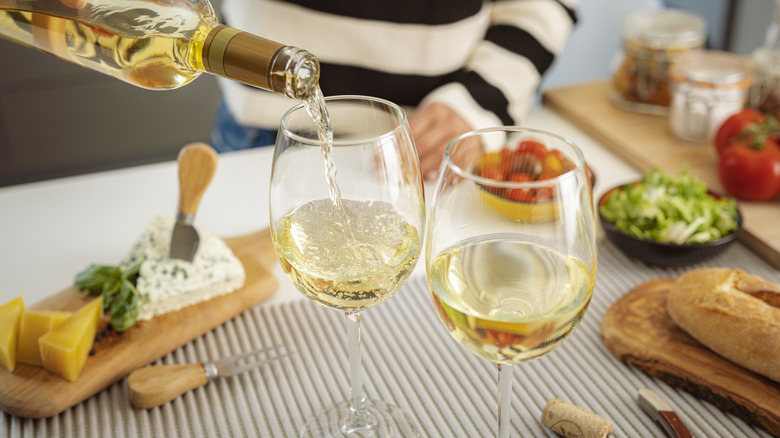 A woman pour two glasses of white wine with a charcuterie spread.