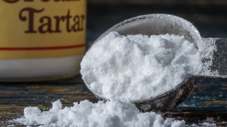 A spoon with overflowing cream of tartar on a dark surface with a jar in the background
