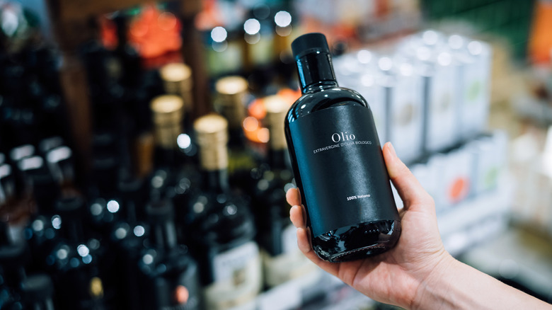 A person stands in a grocery store aisle holding a bottle of expensive-looking olive oil.