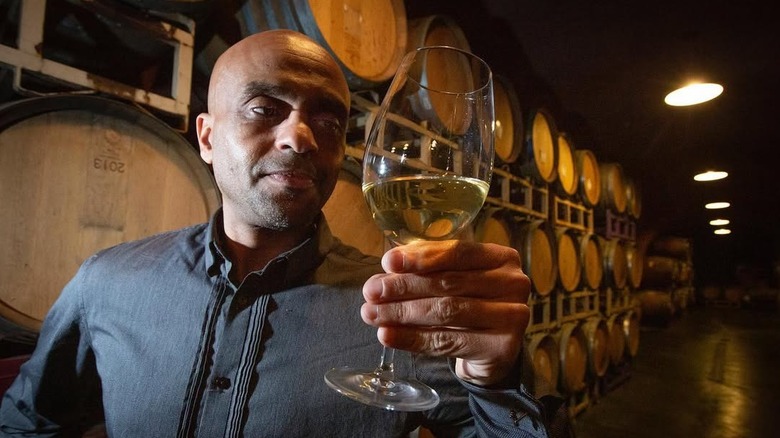 Bee D'Vine founder Ayele Solomon holding a glass of hone wine in front of aging barrels