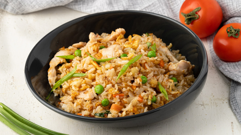 A black bowl of chicken fried rice with green onions and peas inside and some Tomatoes sitting off to the side on the counter