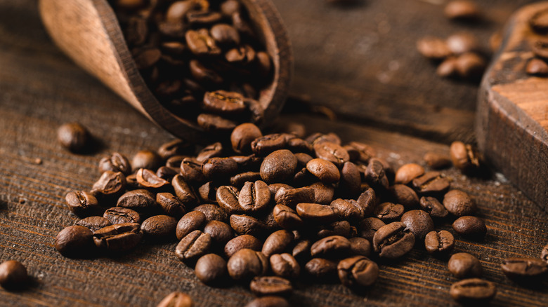a pile of coffee beans on a wooden table with a wooden scoop behind it