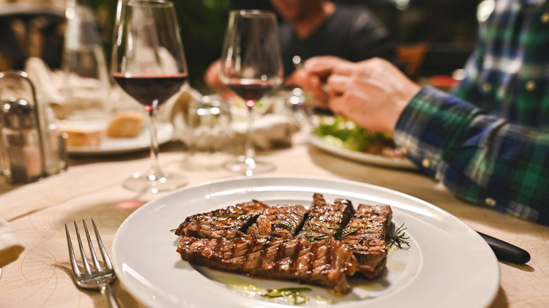 Closeup of a plate of steak at a fancy steakhouse, with diners and wine glasses in the background