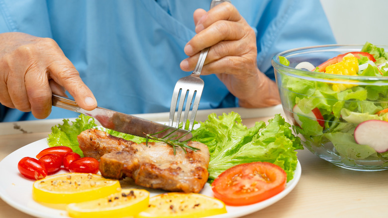 A person in hospital scrubs eating a pork chop on a plate with some lettuce, tomato, and lemon slices next to a side salad