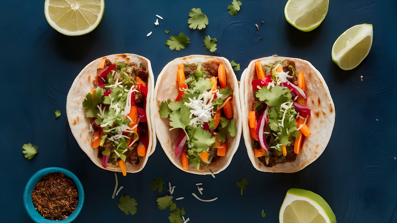 A trio of fresh looking Tacos on a blue background. The tacos are filled with meat and vegetables and topped with cilantro and cheese. There are she shreds and cilantro leaves on the table around them