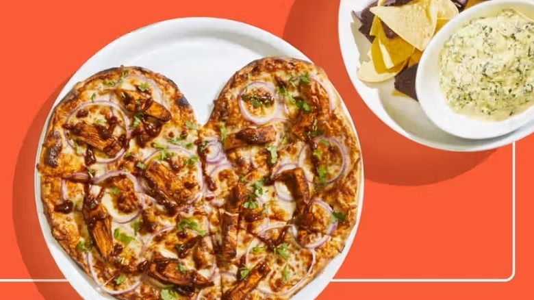 A heart-shaped Pizza on a plate next to a plate of tortilla chips and dip on an orange background