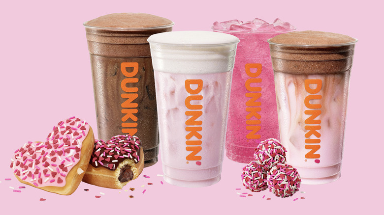 four dunkin' donuts drinks alongside some heart-shaped donuts and sprinkle-covered munchkins on a pink background