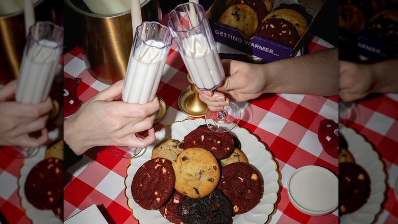 two people clinking milk glasses over a plate of cookies on a table with a red checkered tablecloth