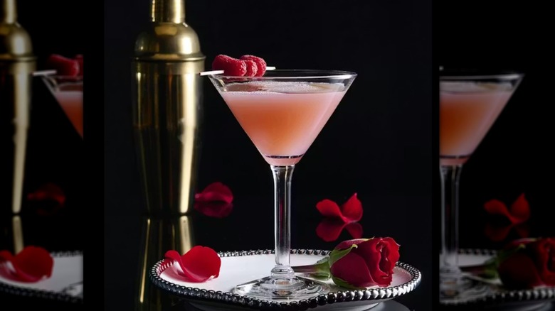A peach colored cocktail garnished with raspberries in a martini glass on a plate with a rose