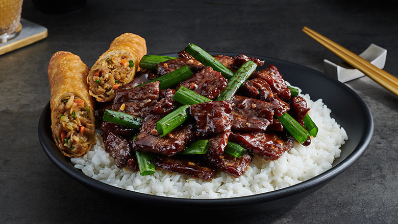 Please beef with green onions on a bed of rice and a black plate with a spring roll on the side