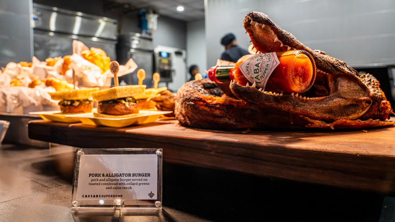 A presentation of pork and alligator burgers at Caesars Superdome featuring an alligator head with a bottle of Tabasco sauce in its mouth