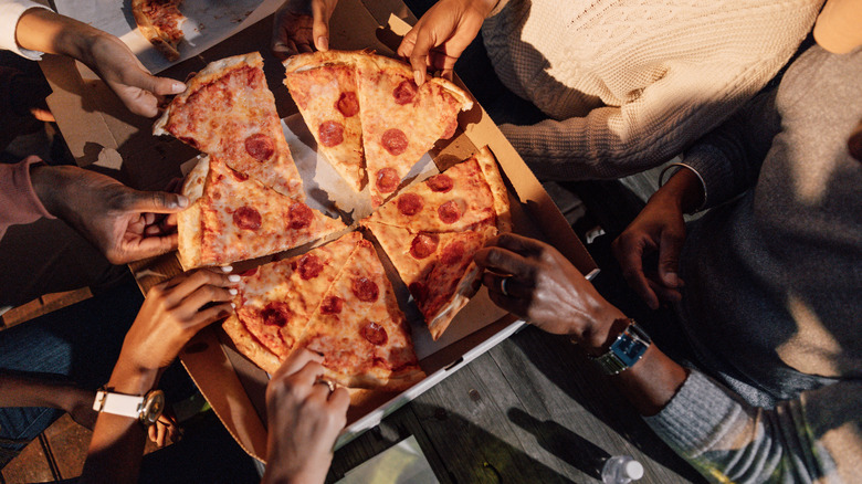 A group of people sitting around a box of pizza reaching for and taking a slice.