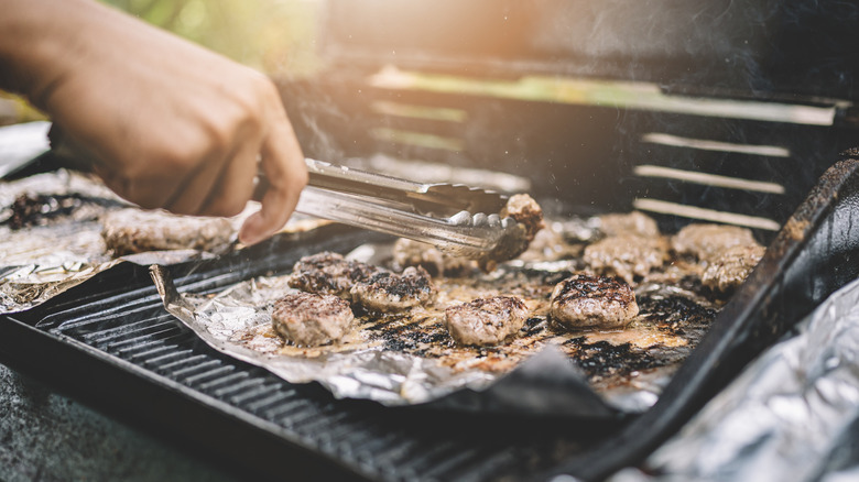 Hand holding tongs moving meat on a grill covered with aluminum foil