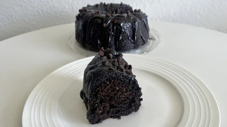 A slice of dark chocolate cake on a white plate in the foreground with the whole cake in the background