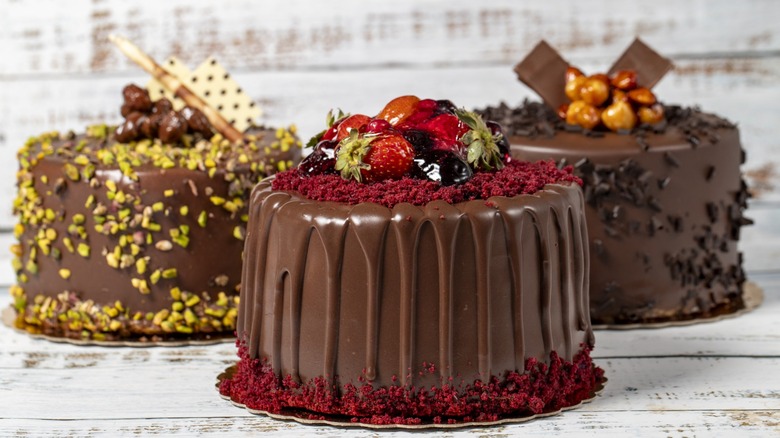 Three chocolate cakes displayed on a table, each decorated differently. One is decorated with fruit, one with pistachio, one with chocolate shavings