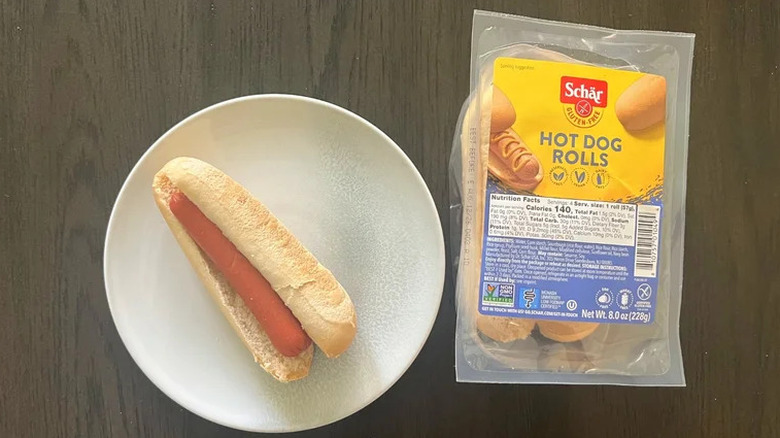 A hot dog inside of a Schär Hot Dog Roll on a white plate, sitting next to a packet of Schär Hot Dog Rolls on a brown wooden table