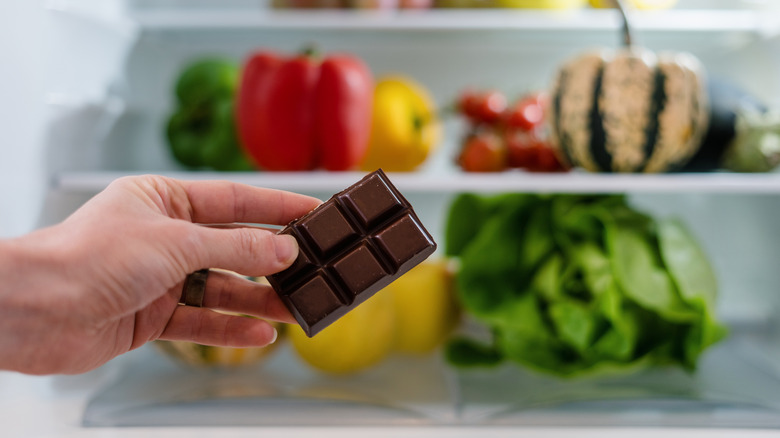 A hand holding a chunk of a chocolate bar in front of an open refrigerator stocked with veggies