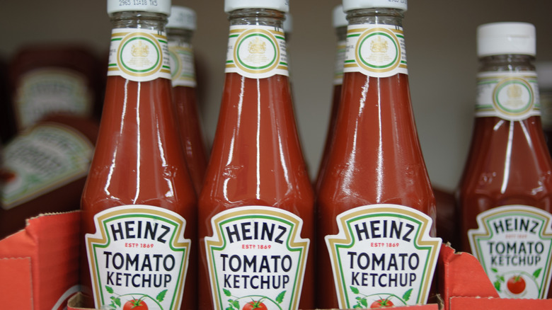 Several glass bottles of Heinz tomato ketchup standing, with more bottles piled behind them