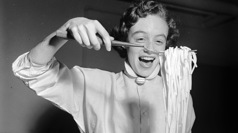 A black and white photograph of a smiling young woman lifting a pile of cooked spaghetti with a metal cooking utensil