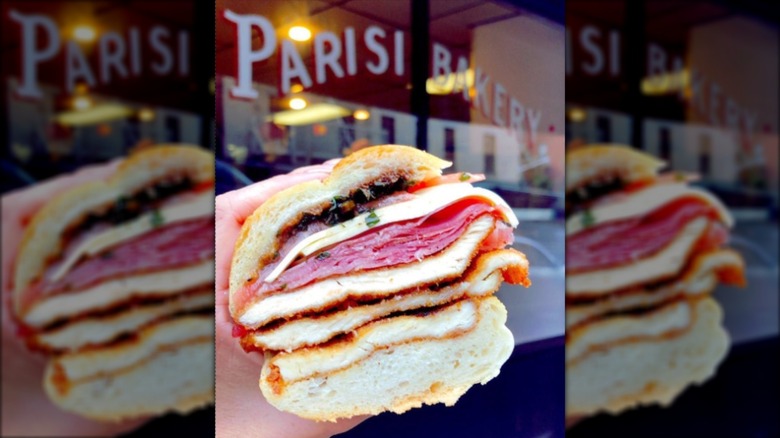 Hand holding the Dennis sandwich in front of a deli's front window