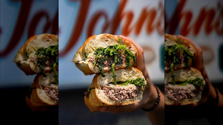 Hand holding two halves of a roast beef sub in front of a John's Deli sign