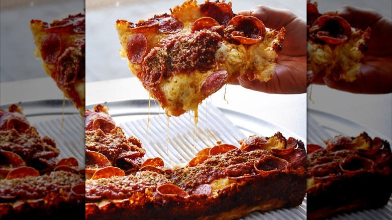 Hand holding one piece of Detroit-style pizza above the rest of the pie