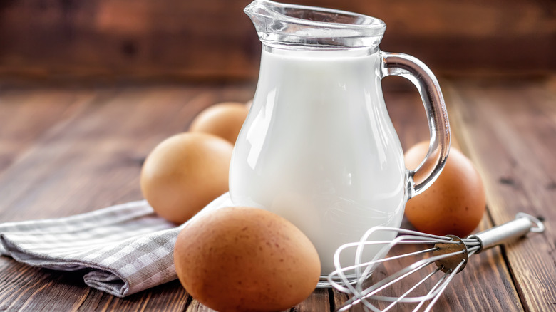 glass pitcher of milk with eggs and a whisk nearby