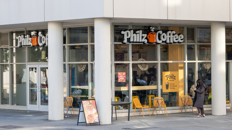 The exterior of a Philz Coffee is pictured with a customer standing beside a pillar, and chairs surrounding the side of the store.