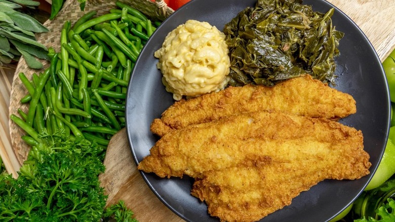 Fried fish filets on plate with mac and cheese and collard greens with fresh veg and greens in background