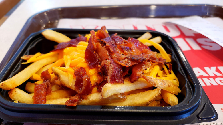 An order of Wendy's Baconator fries in a black plastic container. The french fries are topped with shredded cheese and crumbled bacon