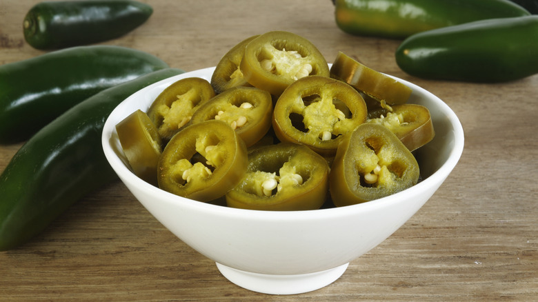 A white bowl full of pickled jalapeno slices with fresh jalapenos in the background