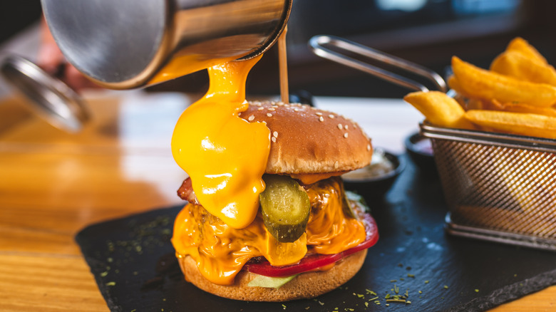 Liquid cheese sauce being poured over the top of a large hamburger with some french fries in the background
