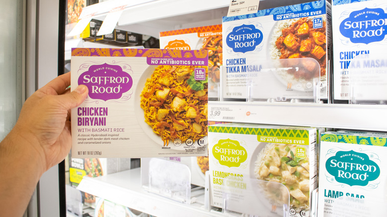 Hand pulling Saffron Road chicken biryani from grocery store freezer case lined with other Saffron Road frozen meals