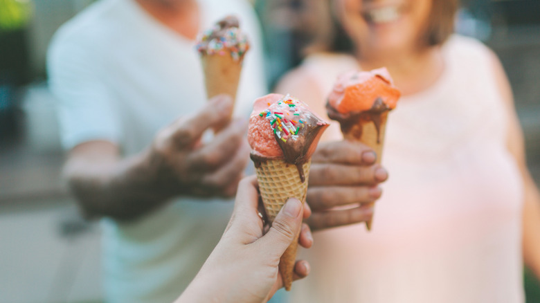 Three hands holding cones of ice cream toped with sprinkles and chocolate sauce