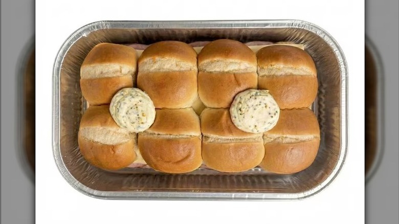 Member's Mark Uncured Ham and Cheese Sliders with pats of herbed butter on top, sitting in an aluminum tin on a white background