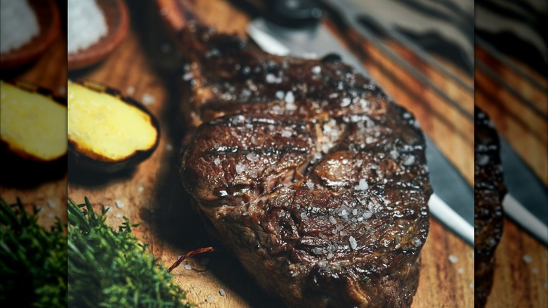 A grilled and salted tomahawk steak on wooden cutting board next to a potato and some herbs