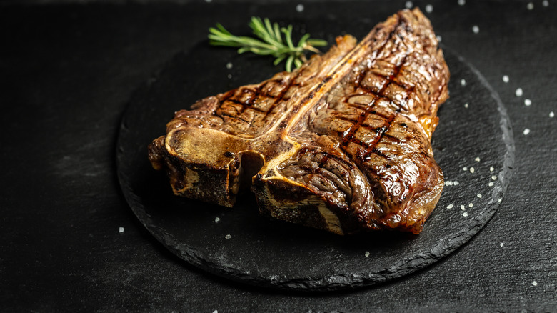 A grilled porterhouse steak on a black stone sprinkled with salt and a sprig of rosemary