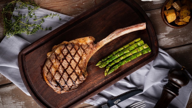 A grilled tomahawk steak on a wooden board next to some grilled asparagus