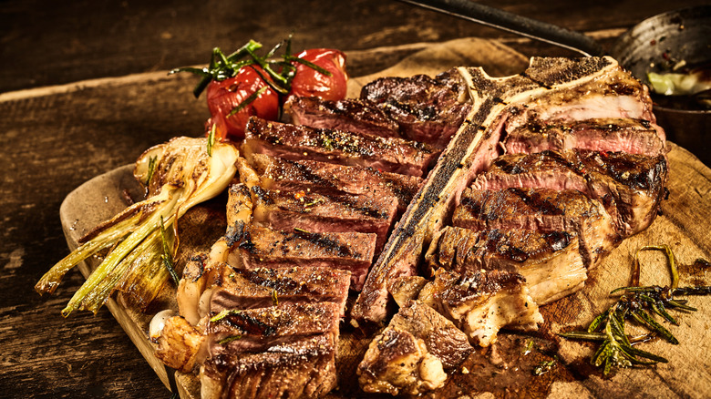 Grilled and sliced porterhouse steak on a wooden board next to some fennel and herbs