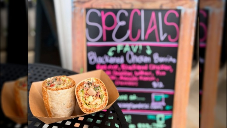 Two halves of a burrito in a cardboard tray in front of menu board