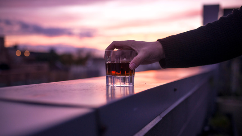 A man holding a tumbler of premium whiskey on a rooftop bar during a colorful sunset.