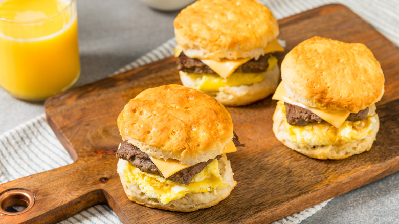 three sausage, egg, and cheese biscuits on a wooden cutting board with a glass of orange juice beside it