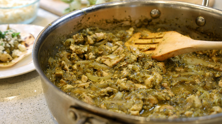 a stainless steel pan full of chicken and onions in verde sauce with a wooden spatula in it