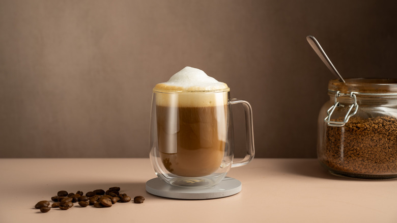 A frothy cup of coffee in a glass mug on a table beside a jar of instant coffee and some fresh coffee beans
