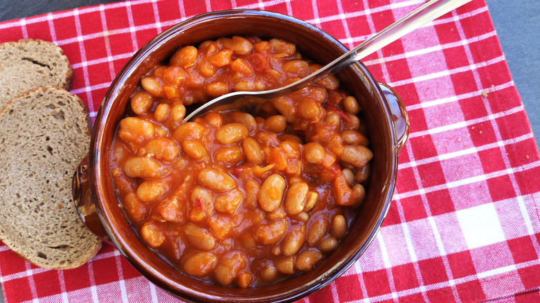 Baked beans in tomato sauce, in a rustic ceramic bowl with whole-grain bread served alongside, on a red and white placemat